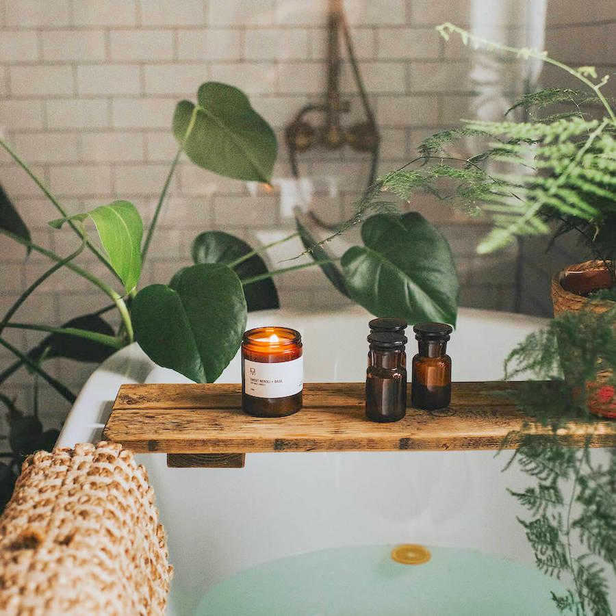 Handmade Candle Co's amber glass sustainable candles, pictured on a wooden plinth on a bath in a white tiled bathroom full of plants