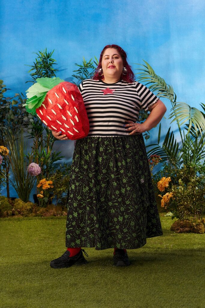 Person in a black and white stripe t-shirt and a floral skirt from sustainable clothing brand Lucy & Yak. They're in a garden holding a giant strawberry made from paper.